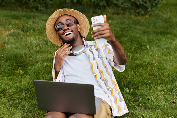 A joyful young man relaxes outdoors, working remotely with his laptop and smartphone in hand.