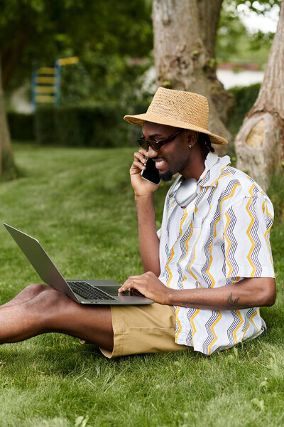 A young African American man works on his laptop and chats on the phone in a lush green park.