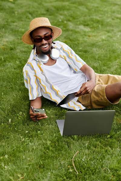 Young man relaxes on grass, working remotely with a laptop and smartphone in a sunny environment.