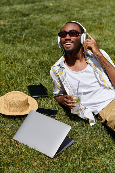 A young african american man relaxes on the grass, wearing headphones and working on his laptop.