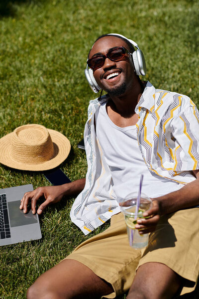 A young man relaxes on grass, working on his laptop while enjoying a refreshing drink and sun.