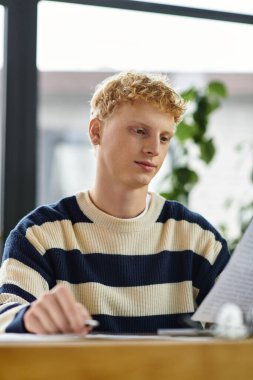 Çizgili kazaklı genç bir kızıl adam çağdaş bir ofiste evrak işlerine odaklanıyor..