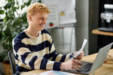Genç kızıl saçlı bir adam, parlak bir ofiste belgeleri incelerken dizüstü bilgisayarla meşgul oluyor..