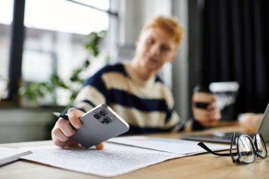 Çizgili kazaklı genç adam ofiste akıllı telefon kullanırken belgeleri gözden geçiriyor..