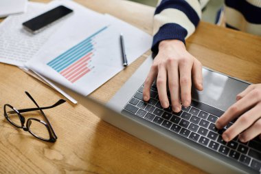 A young redhead man types on a laptop while surrounded by office materials and reports. clipart