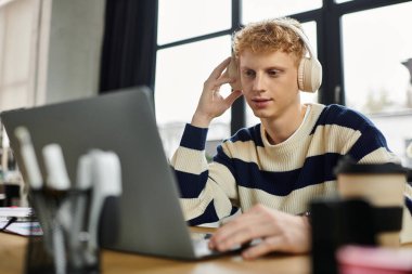 Çizgili kazaklı kızıl saçlı adam canlı bir ofis ortamında dizüstü bilgisayarıyla meşgul oluyor..