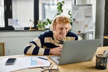 A cheerful young redhead male manager focuses on his laptop while enjoying a drink. clipart