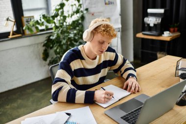 Redhead man in striped sweater concentrates on writing notes while seated at his desk. clipart