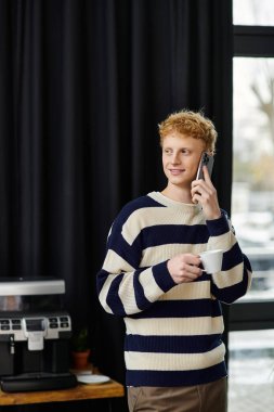 Çizgili kazaklı genç bir kızıl adam elinde bardak tutarken telefonla konuşuyor..