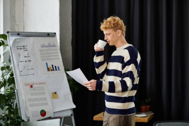 Redhead man in a striped sweater reviews documents while enjoying coffee at his corporate workspace. clipart