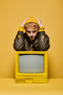 Young redhead man showcases fashionable winter attire leaning on a retro TV against a bright yellow backdrop. clipart