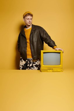 Kış kıyafetli modaya uygun genç bir adam stüdyoda eski bir televizyonla güven içinde poz veriyor..