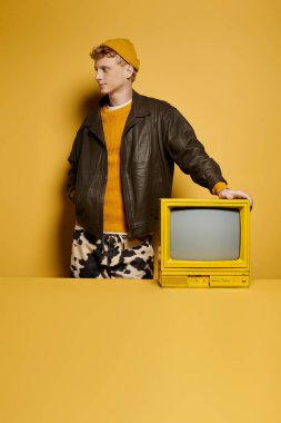 Şık bir kızıl saçlı, modaya uygun bir kış kıyafetinin içinde sarı bir arka plana yaslanmış bir retro televizyona yaslanarak poz veriyor..