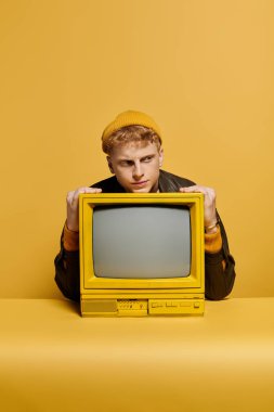 Havalı bir kızıl saçlı adam, canlı sarı bir arka plana karşı retro televizyon sergiliyor..