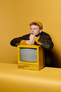 A fashionable young man with red hair relaxes beside a vintage yellow television in a studio. clipart