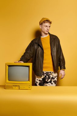 Tarz sahibi genç bir kızıl, sarı bir arka fona karşı klasik bir televizyonla, modaya uygun bir kış elbisesi giymiş olarak poz veriyor..