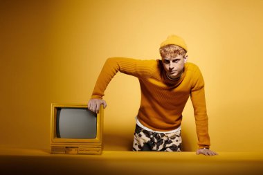 Şık, genç bir kızıl, canlı bir kış elbisesiyle, klasik televizyonun yanında, sarı bir arka fona karşı güven içinde poz veriyor..