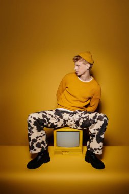 Şık, genç bir kızıl, sarı arka planda klasik bir televizyonun karşısında otururken kendine güveni olan bir kış kıyafetiyle poz veriyor..