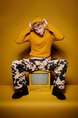 Şık, kızıl saçlı bir genç, şık bir kış elbisesi içinde, klasik bir televizyonun önünde, sarı bir arkaplanda poz veriyor..