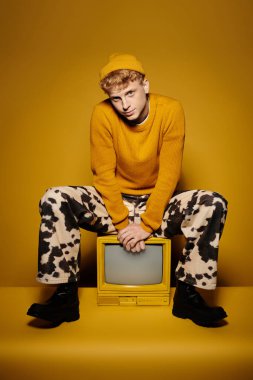 Young man in a vibrant winter outfit sits confidently with a vintage television in a studio. clipart