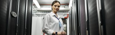 Beyaz gömlekli bir kadın şık bir veri merkezinde sunucu operasyonlarını denetliyor, güvenliği ve pankartı sağlıyor.