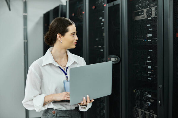 Женщина в белой рубашке усердно работает над ноутбуком в серверной комнате, обеспечивая надежность системы.