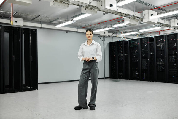 Женщина в белой рубашке стоит в гладкой, современной серверной комнате, сосредоточенная на своих задачах.