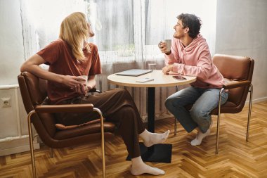 Two loving men sit together in a sunny room, sharing smiles and coffee at a small table. clipart