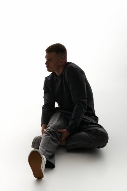 Genç bir adam yerde oturuyor, bacağını nazikçe yumuşak bir ışıkta tutuyor..