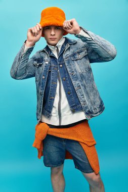 Young man in a denim jacket and bright orange hat poses against a blue backdrop. clipart