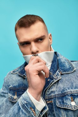 Şık bir genç adam kendine güvenerek poz verir, kot ceket giyer ve beyaz balıkçı yaka yaka giyer..