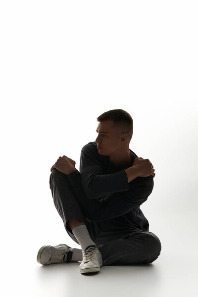 Молодой человек сидит изящно с руками на коленях, потерявшись в мыслях на свежем фоне.