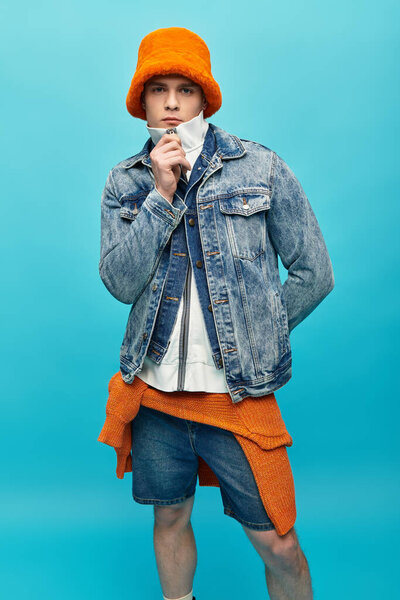 A young man in a denim jacket and orange hat shows off his trendy style.