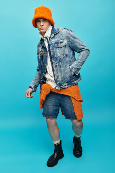 Уверенный молодой человек в джинсовой куртке и оранжевых аксессуарах позирует на ярком фоне.