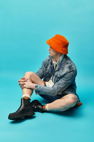 A stylish young man in a denim jacket and orange bucket hat sits cross legged on a soft surface.