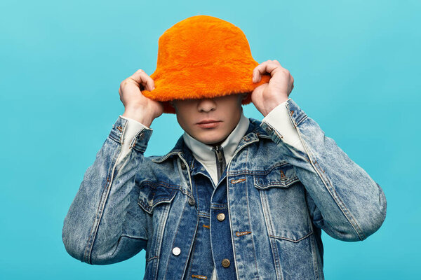 Молодой человек в пушистой оранжевой шляпе улыбается перед ярко-голубой стеной.