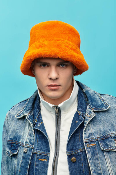 Handsome young man showcases a stylish orange hat, dressed in denim, against a bright blue backdrop.