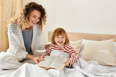 Kıvırcık saçlı anne ve kızı, yatakta okurken, beraberliği kucaklarken keyifli bir anı paylaşıyorlar..