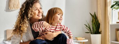 Kıvırcık saçlı bir anne, evinde rahat bir anın tadını çıkarırken gülüyor ve kızını kucaklıyor.