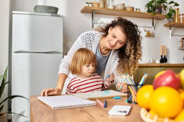 Neşeli bir anne, kızına rahat mutfak masasında yaratıcı bir çizim aktivitesinde rehberlik eder..