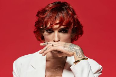 Kızıl saçlı ve dövmeli genç bir adam stilini parlak kırmızı bir arka planda sergiliyor..