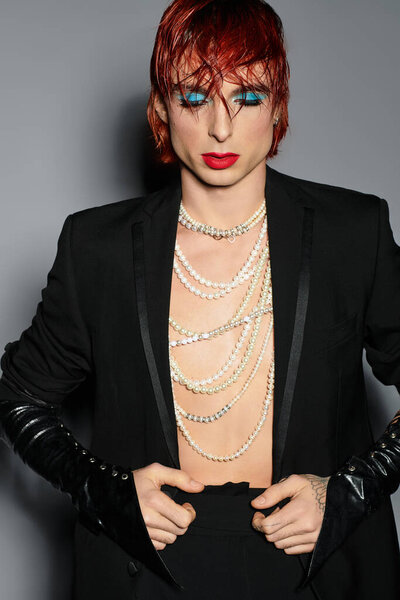 Молодой человек с рыжими волосами носит смелый синий макияж и элегантный наряд в стильной обстановке.