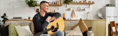Genç bir adam şık bir dairede kanepede dinleniyor gitar çalıyor ve müzikten zevk alıyor.