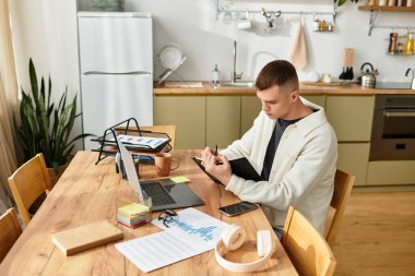 Handsome young man in casual attire sits at a wooden table, focused on work tasks in his apartment. clipart