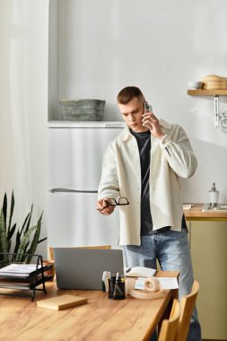 Sıradan giyinmiş odaklanmış genç bir adam, elinde telefon ve gözlük tutarken uzak bir işle meşgul oluyor..