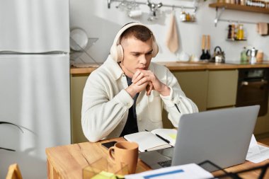 A young man in casual attire thoughtfully works on his laptop in a stylish home setting. clipart