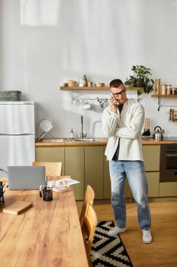 Gündelik giysiler içindeki genç bir adam modern bir dairede telefonuyla konuşuyor.