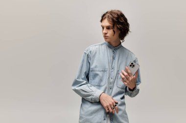 Yakışıklı bir genç adam şık bir kot gömlekle düşünceli bir şekilde poz veriyor, elinde akıllı bir telefon var..