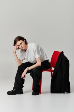 Şık görünümlü genç bir adam, çağdaş bir stüdyoda kırmızı bir sandalyede düşünceli bir şekilde oturur..