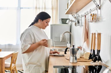 Güzel genç bir kadın güneşli bir mutfakta durup telefonuna bakarken kahvenin tadını çıkarıyor..